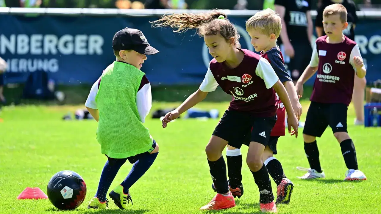 Kinder im Mannschaftssport Fußball üben auf einem grünen Rasen. Ein Junge mit grüner Weste dribbelt den Ball, während ein Mädchen und ein weiterer Junge ihn beobachten.