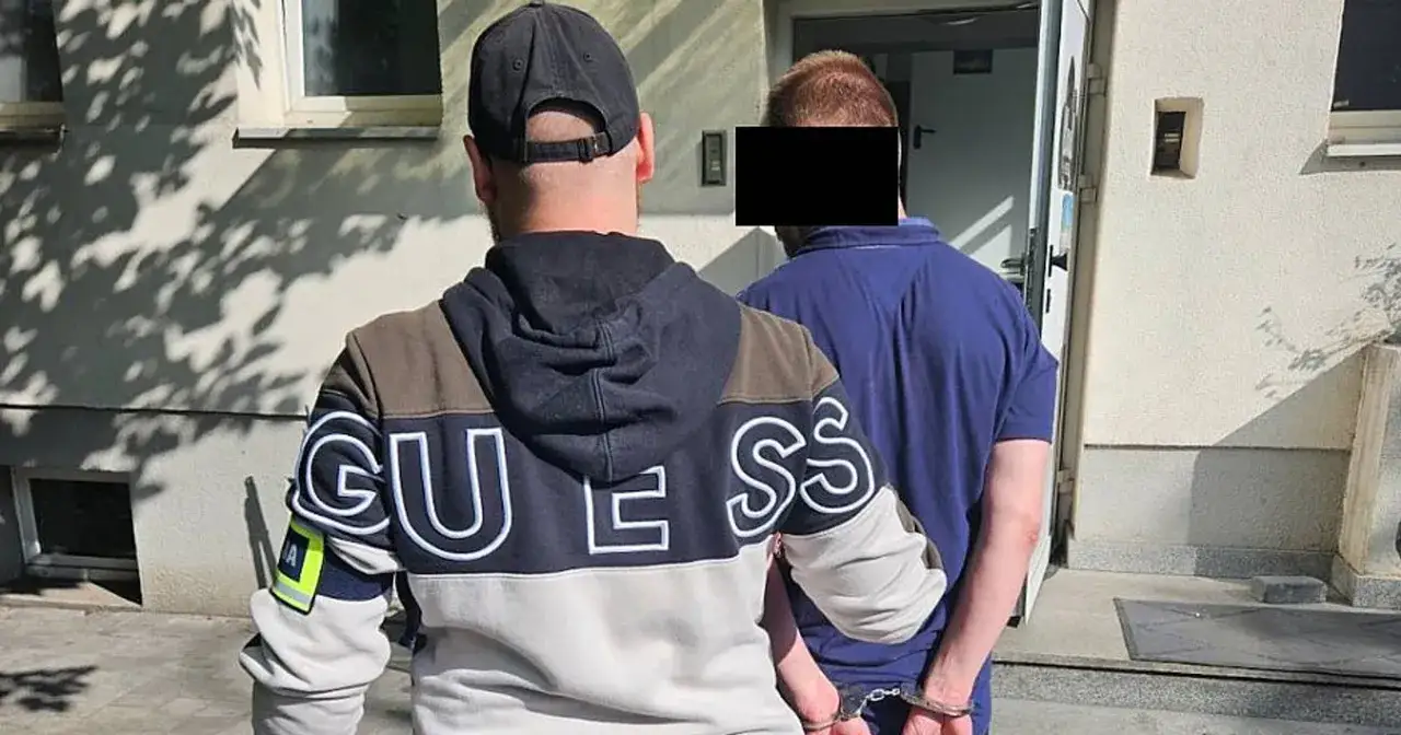 Policjant w kajdankach prowadzi zatrzymanego. Widać przesłanki do zatrzymania, gdy mężczyzna w niebieskiej koszulce jest wyprowadzany.