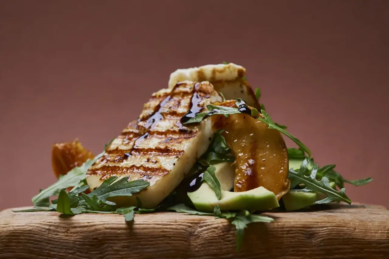 Tranches de halloumi grillé, arrosées de sauce, sur un lit de roquette et d'avocat, avec des morceaux de fruits caramélisés.