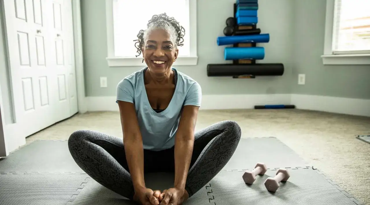 Femme souriante en tenue de sport, assise en tailleur sur un tapis de gym. Des haltères roses sont à côté. Pas de chaise visible.