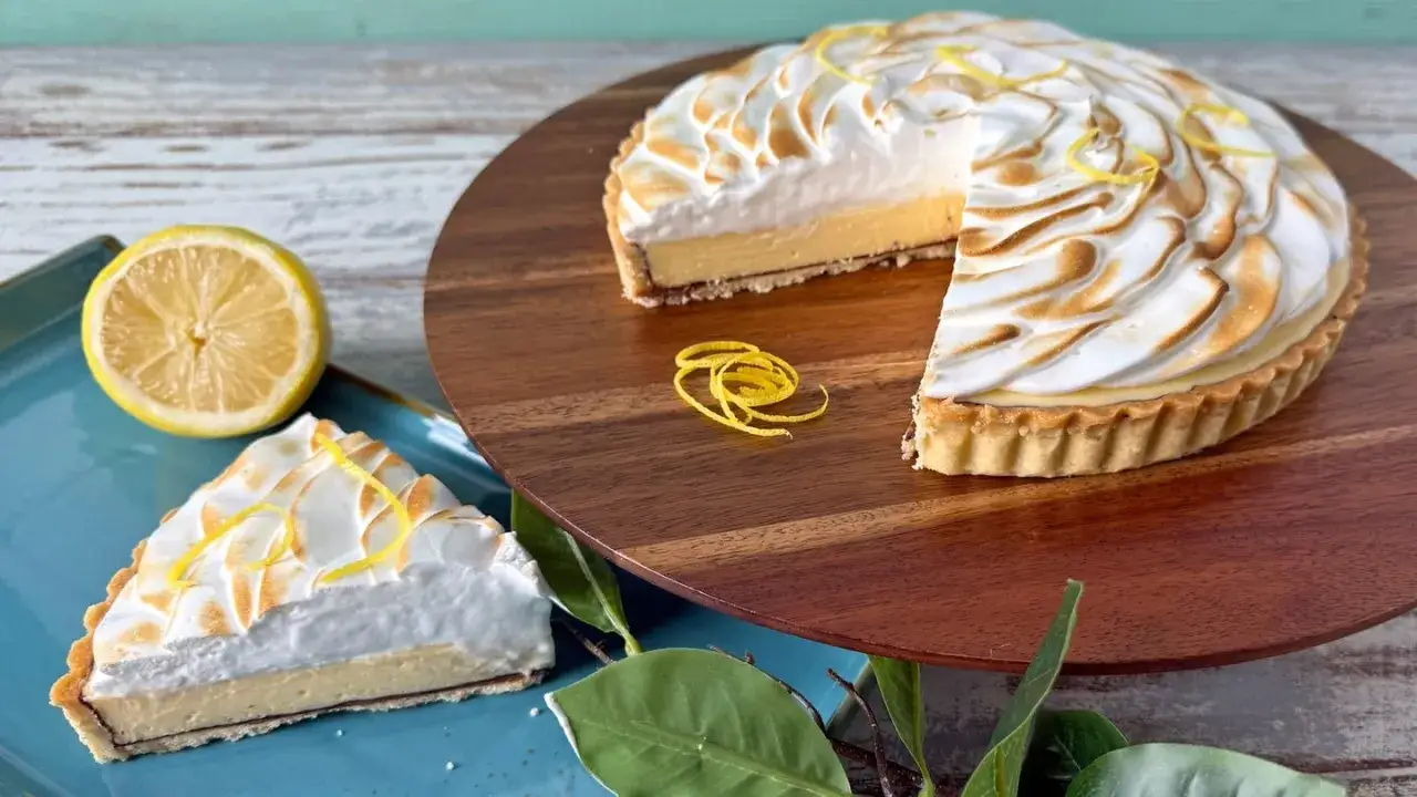 Ein k&ouml;stliches Zitronentarte Rezept: Ganze Tarte mit Baiserhaube und ein St&uuml;ck auf blauem Teller, garniert mit Zitronenzesten.