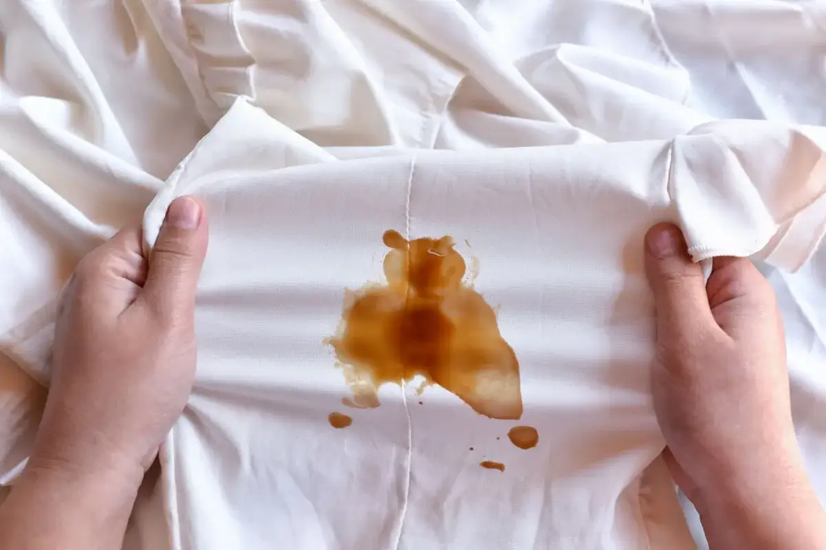 Mani che mostrano un vestito bianco macchiato di caffè, forse caduto da una borsa in pelle.
