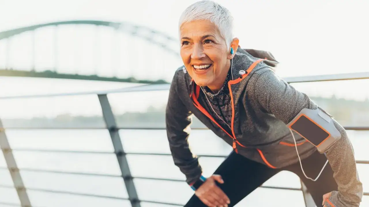 Femme de 70 ans souriante, prête à battre son record de 10 km. Elle porte des écouteurs et un brassard pour son téléphone.