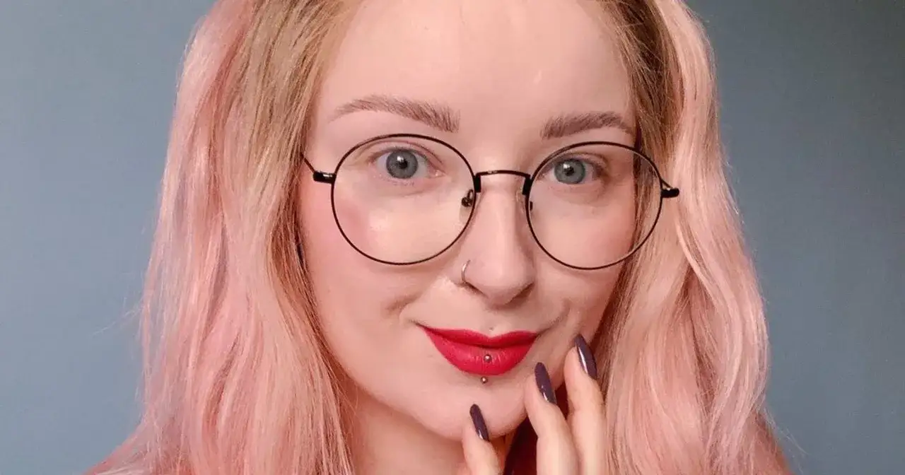 Uśmiechnięta, młoda influencerka z różowymi włosami, okularami i makijażem.