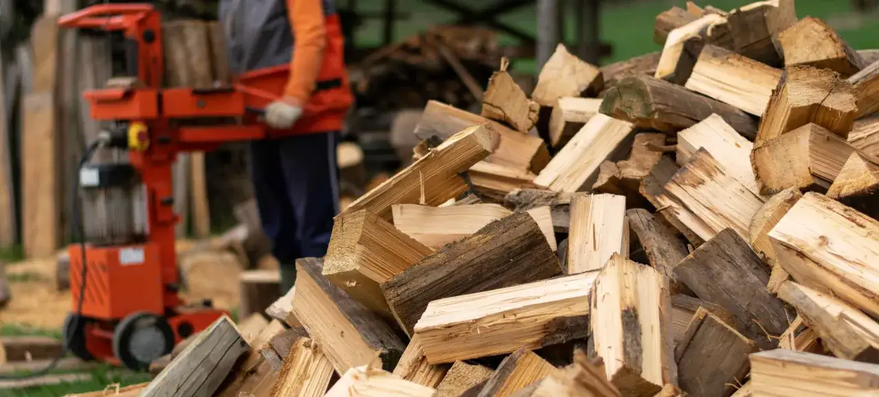 Ein Mann bedient einen roten Holzspalter mit Einhandbedienung, daneben liegt ein großer Haufen gespaltenes Holz.