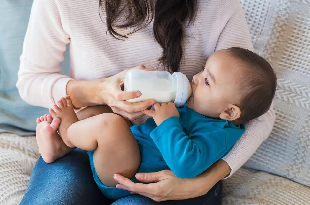 Malutki, 3-tygodniowy niemowlak w niebieskim body pije mleko z butelki, trzymany na rękach.