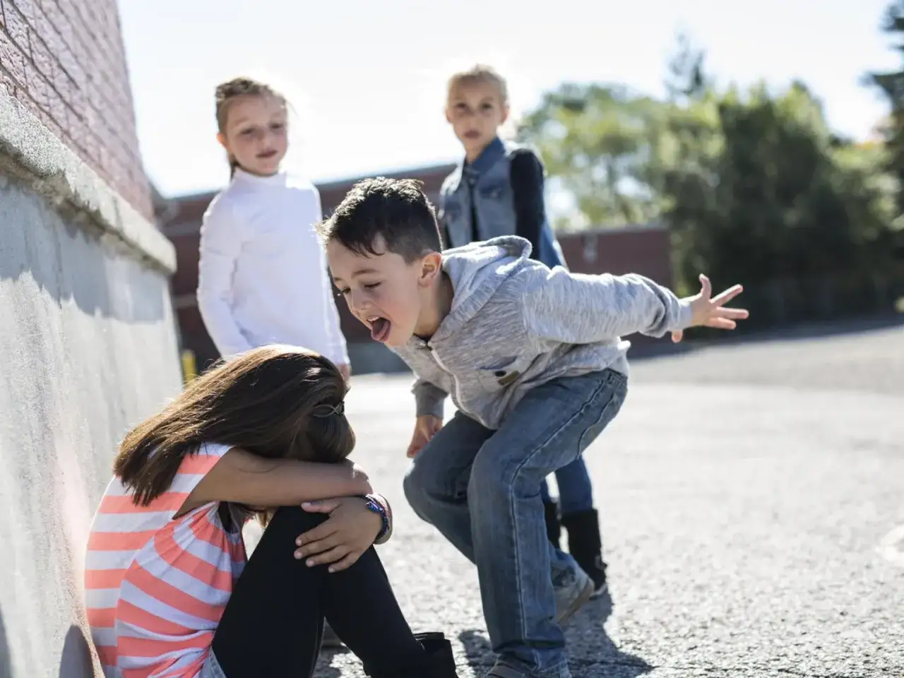 Ein Junge schreit ein Mädchen an, das sich am Boden kauert. Andere Kinder schauen zu. Ein trauriges Bild von Mobbing in der Grundschule.