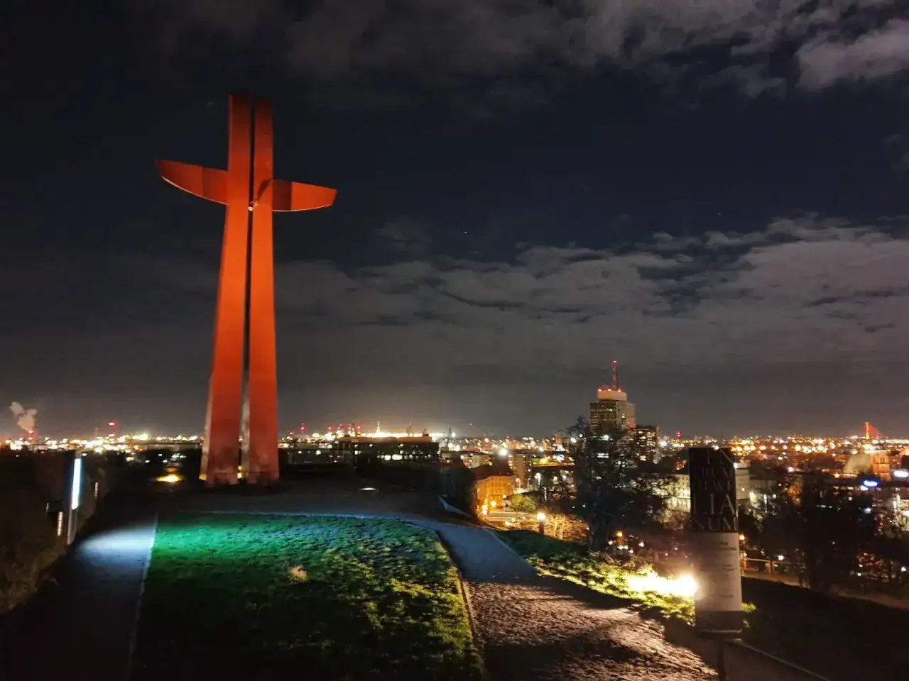 Pomnik na G&oacute;rze Gradowej nocą, oświetlony na pomarańczowo, z widokiem na rozświetlone miasto w tle.