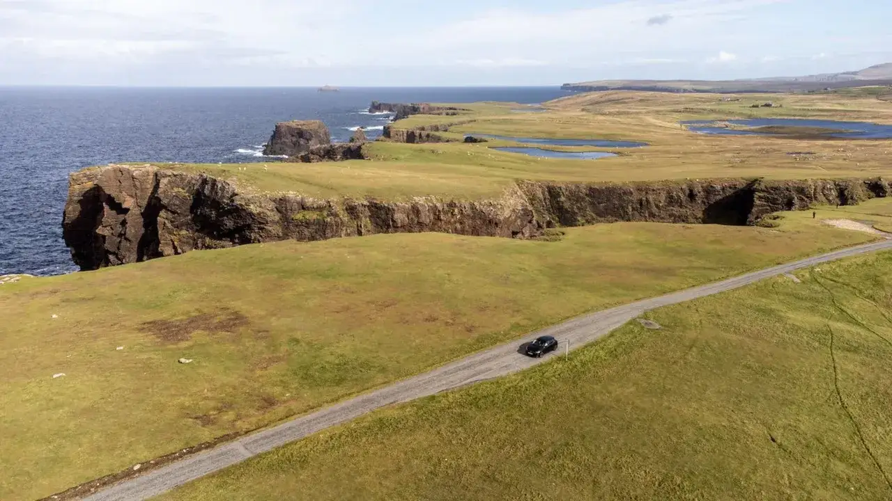 Ein schwarzes Auto f&auml;hrt auf einer einsamen Stra&szlig;e entlang der dramatischen Klippen der Shetlandinseln. Ein perfekter Schauplatz f&uuml;r einen Shetland Krimi.