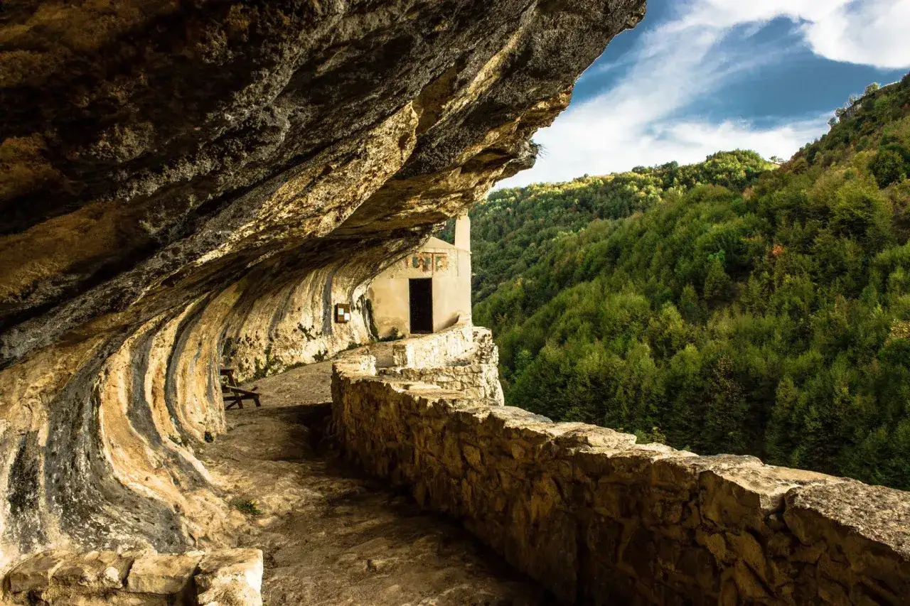 Un sentiero roccioso conduce a un eremo scavato nella roccia, tra i boschi rigogliosi dell'Abruzzo. Un luogo perfetto per i cammini.