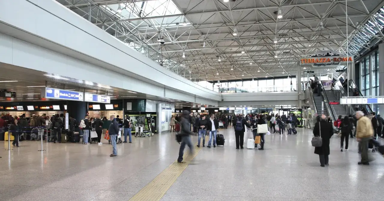 Tłoczny terminal lotniska Rzym Ciampino. Pasażerowie z bagażami czekają w kolejce do odprawy, inni idą w kierunku schodów ruchomych.