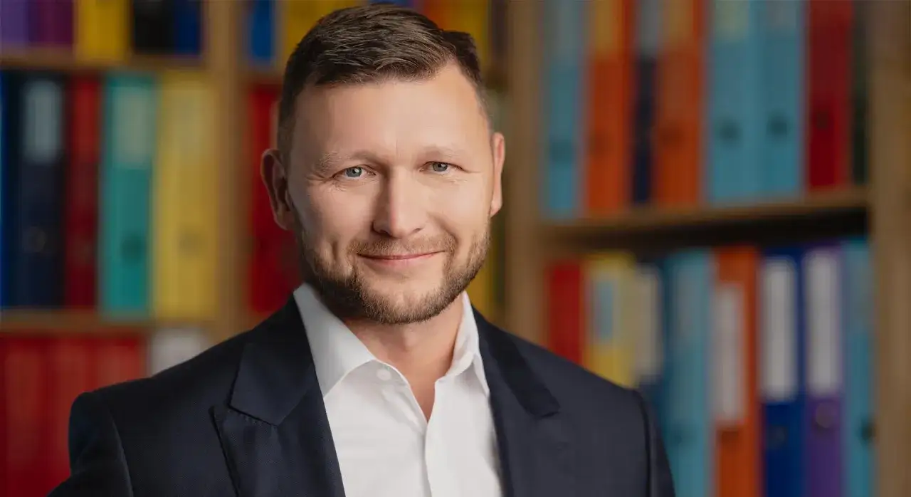 Portret mężczyzny w garniturze na tle kolorowych segregatorów.