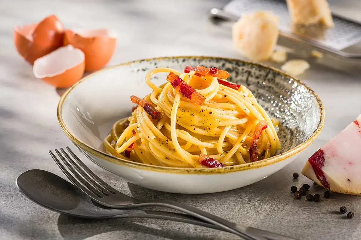 Eine cremige Carbonara nur mit Eigelb, garniert mit knusprigem Speck und frisch gemahlenem Pfeffer. Daneben liegen Eierschalen und ein Stück Pecorino.