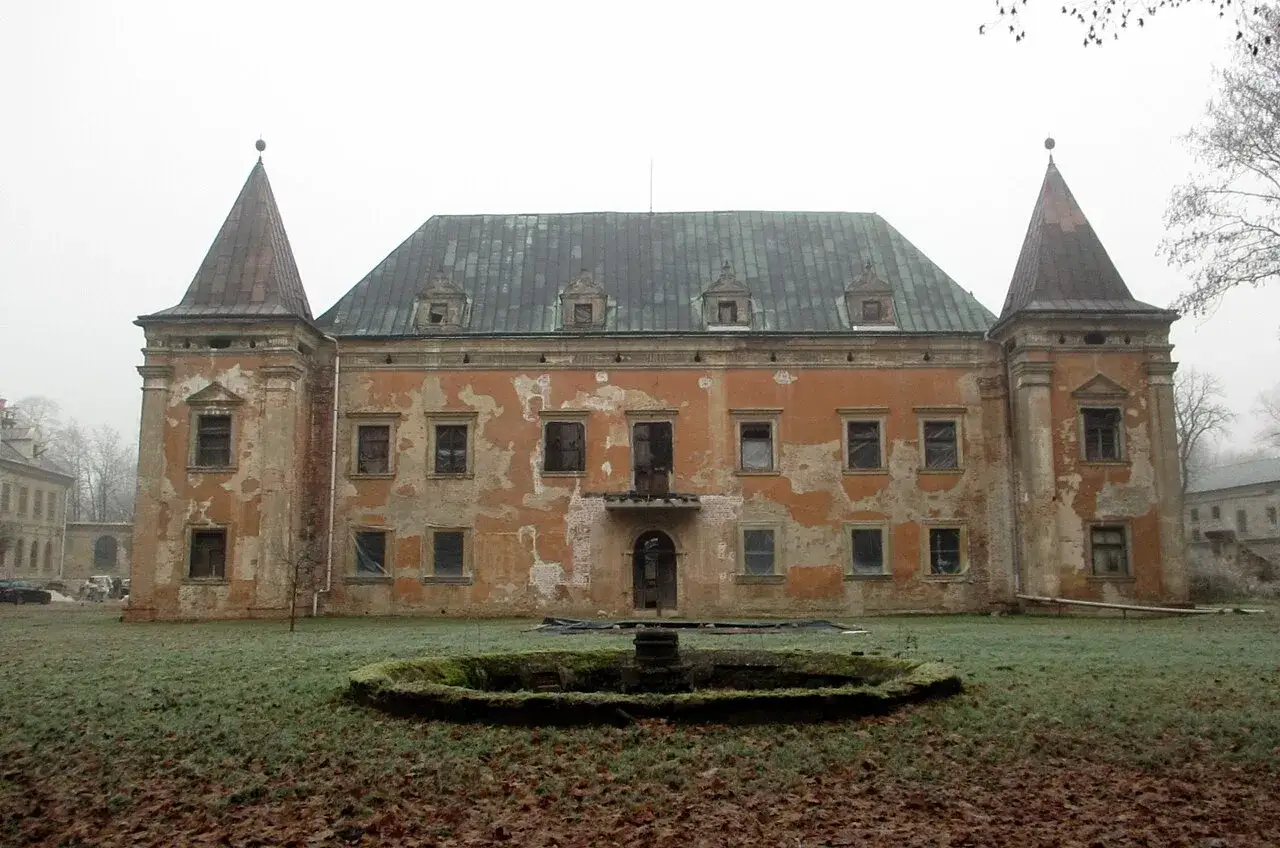 Zrujnowany pałac z wieżyczkami i opadającą tynkiem fasadą, otoczony zaniedbanym ogrodem. Kruszyna historii w mglisty dzień.