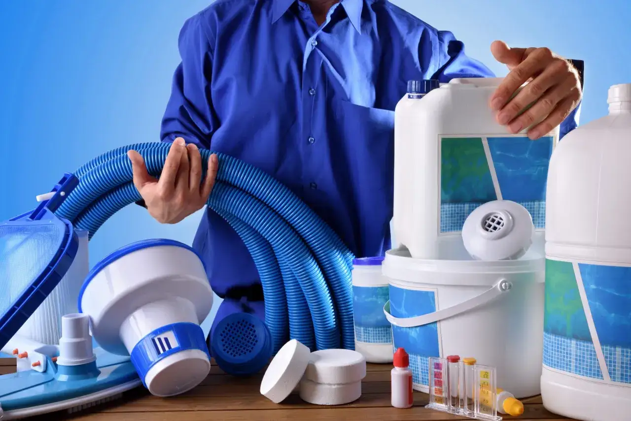 A person in a blue shirt holds a blue vacuum hose, surrounded by pool cleaning supplies. This image shows how to vacuum a pool.