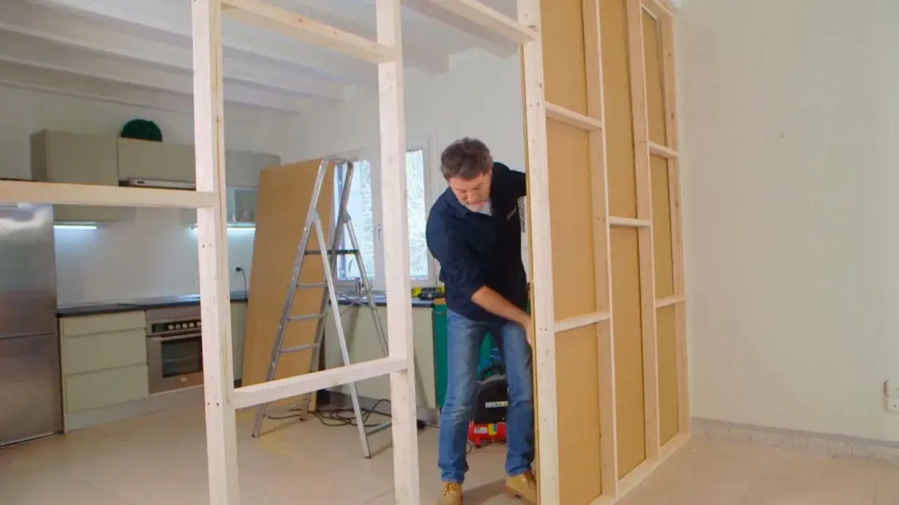 Hombre trabajando para hacer pared de madera. La estructura de madera se ve en primer plano, con una cocina al fondo.