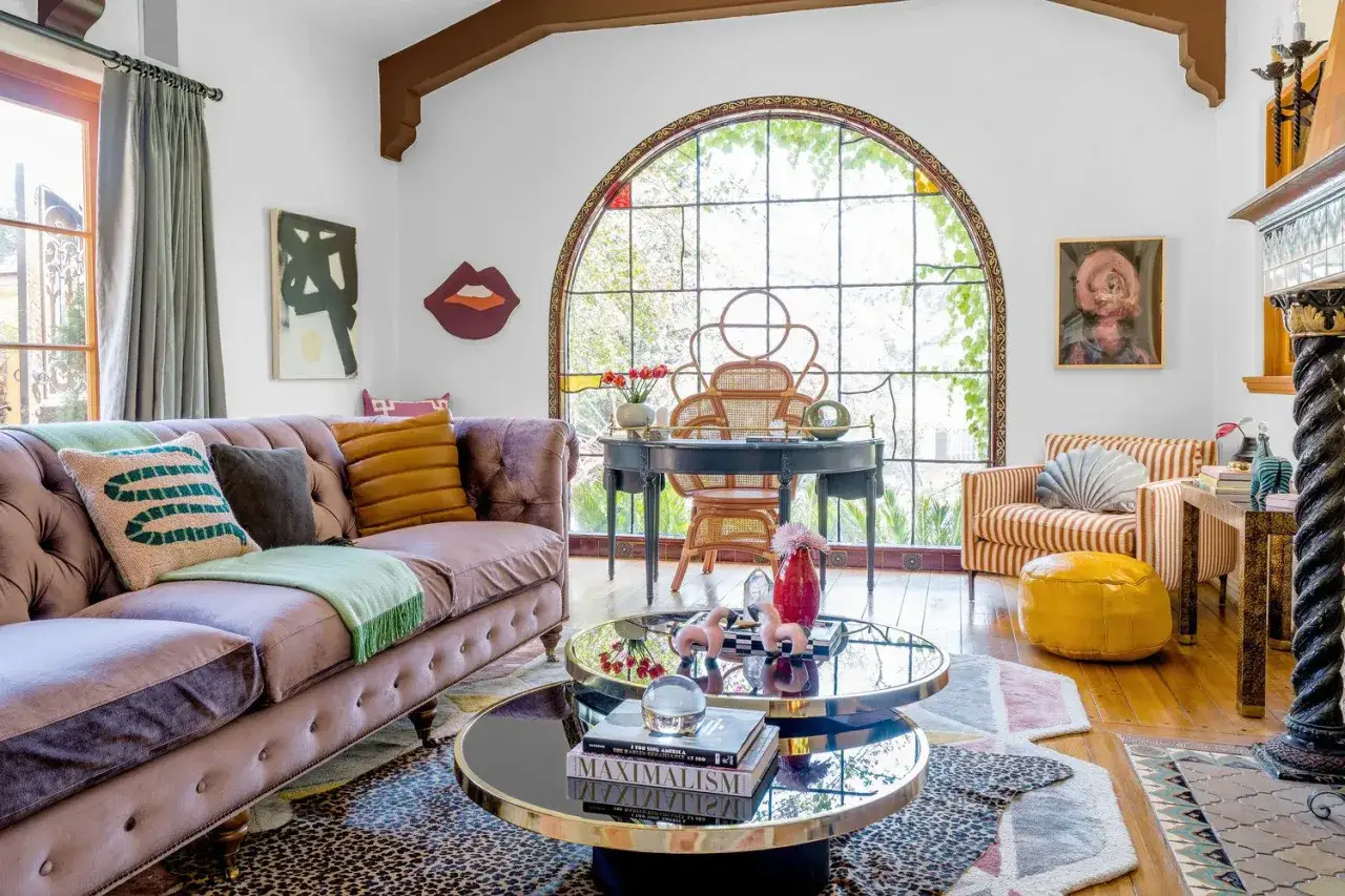Salon w stylu Memphis: fioletowa sofa, stoliki kawowe z lustrzanymi blatami, kolorowe poduszki i dywany, krzesło z rattanu przy biurku pod łukowym oknem.