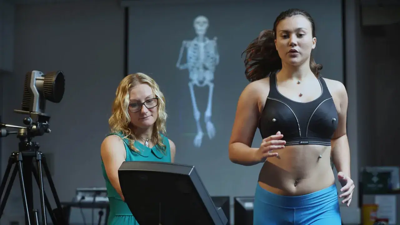 A woman runs on a treadmill wearing a sports bra with sensors, as a researcher observes. A projected skeleton appears behind them.