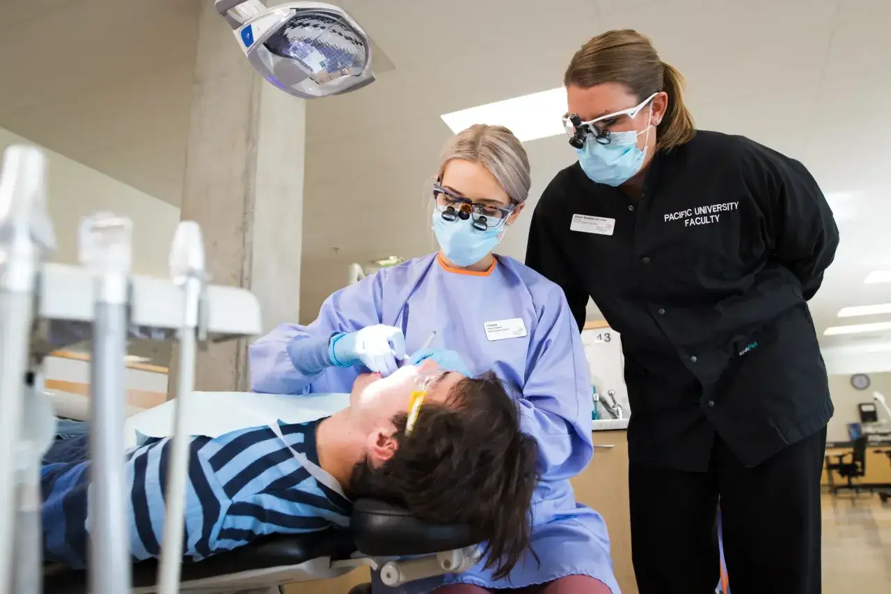Dwie higienistki stomatologiczne w maseczkach i okularach pracują przy pacjencie. Zastanawiasz się, ile zarabia higienistka stomatologiczna?