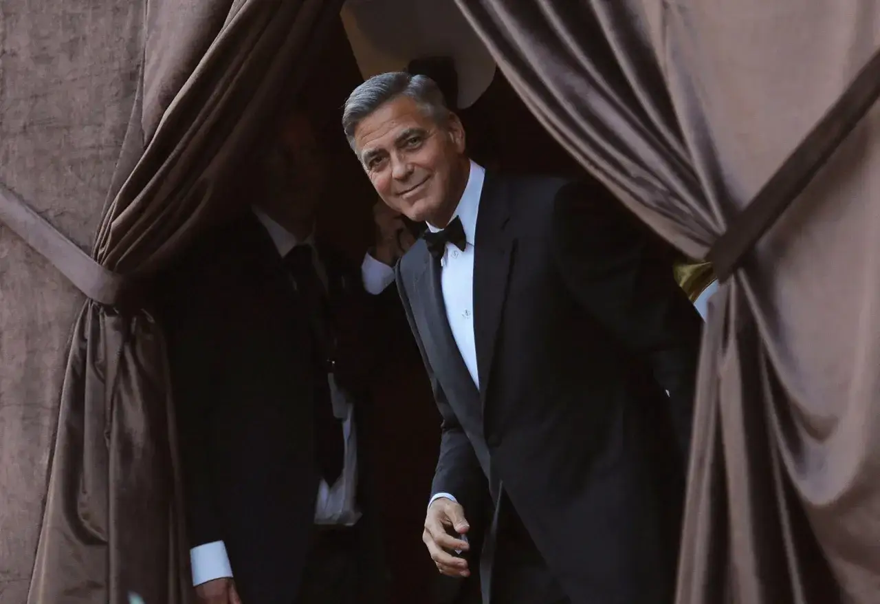 George Clooney, embodying the black tie meaning, smiles as he emerges from behind draped curtains in a classic tuxedo.