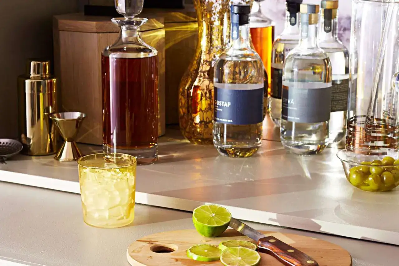 Na blacie stoją **podstawowe alkohole do drink&oacute;w**: whisky, w&oacute;dka i gin. Obok szklanka z lodem, limonka i oliwki.