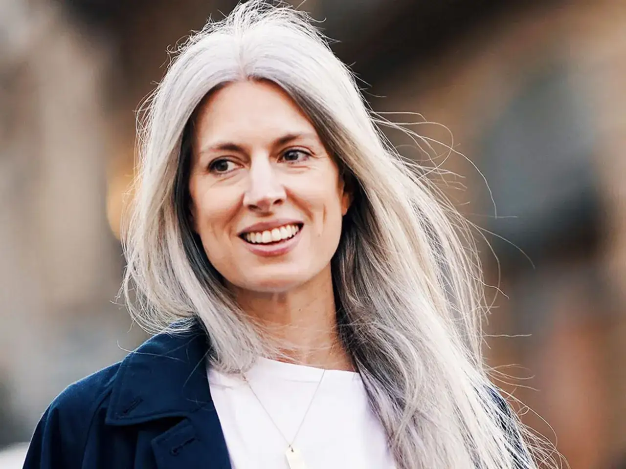 Mujer sonríe con su pelo con canas y mechas rubias, luciendo un abrigo azul marino.
