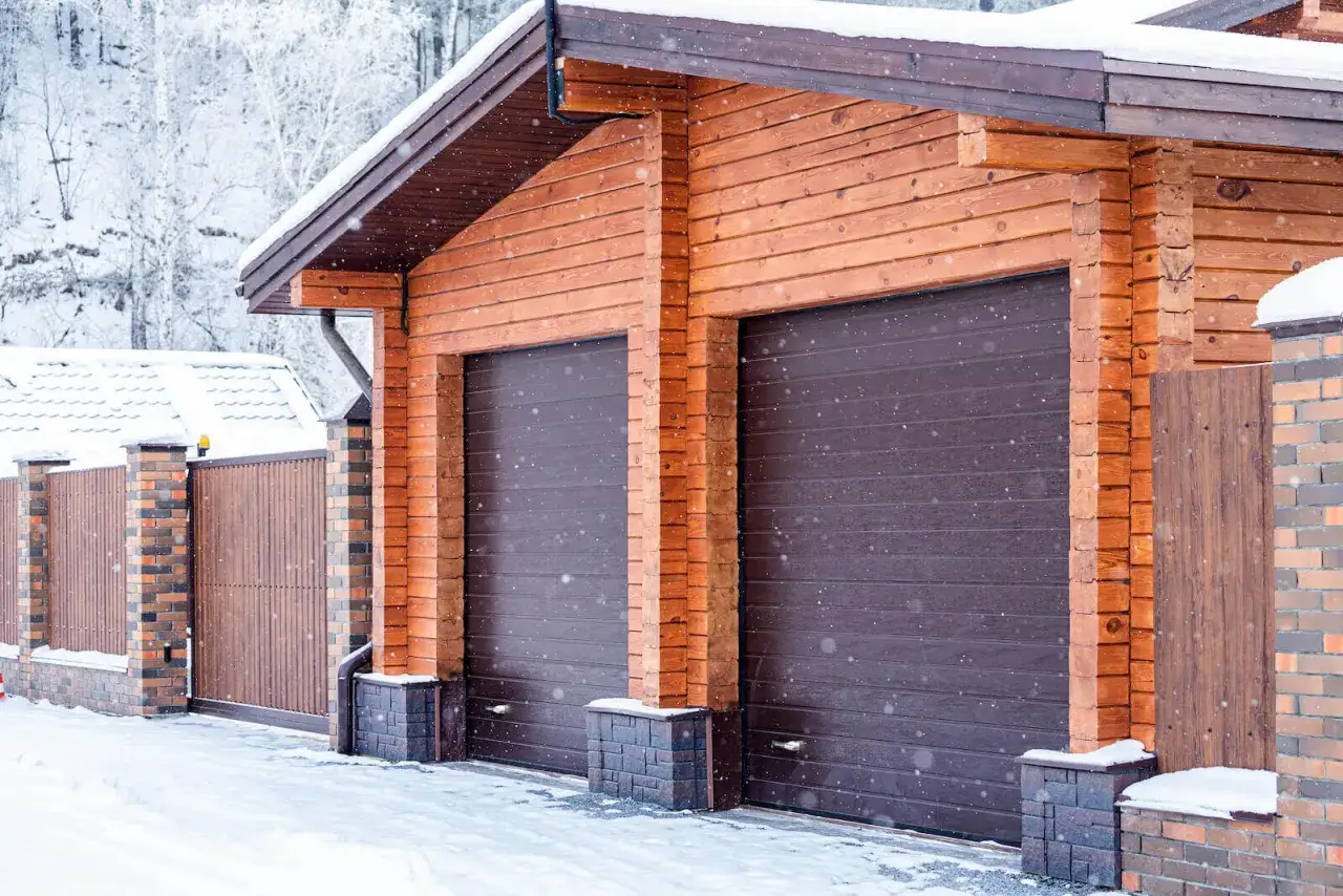 Drewniany garaż z dwoma ciemnobrązowymi bramami, stojący 1,5 m od granicy. Wokół śnieg i padający puch.
