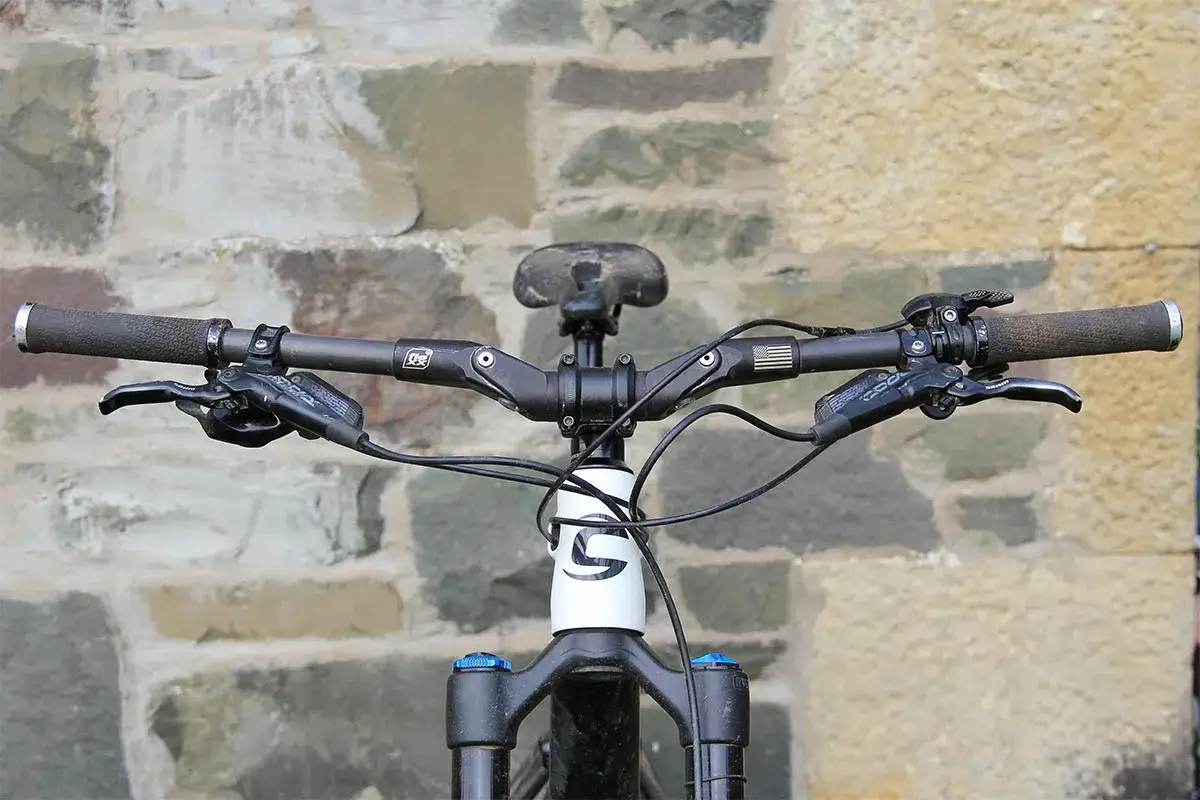 Close-up of a mountain bike's handlebars, showing textured grip damper, brake levers, and a white frame with a black logo.