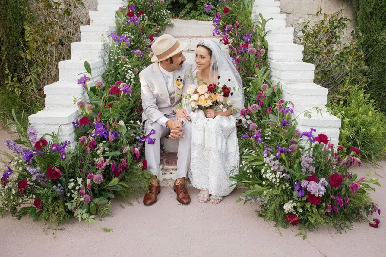 Pareja sonriente sentada en escalones blancos rodeados de flores. Ella lleva velo y vestido de encaje, &eacute;l sombrero y traje. &iquest;Es una boda, no boda?