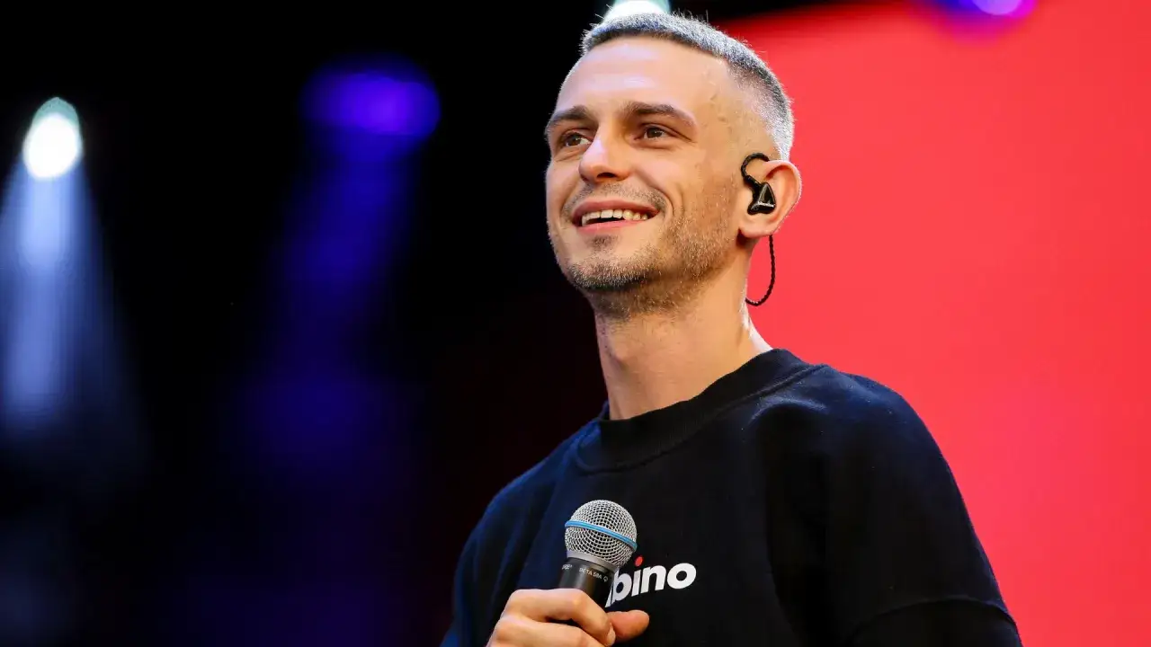 Vito Bambino z mikrofonem, uśmiechnięty na tle czerwonej sceny. Zapowiada swoją nową płytę.