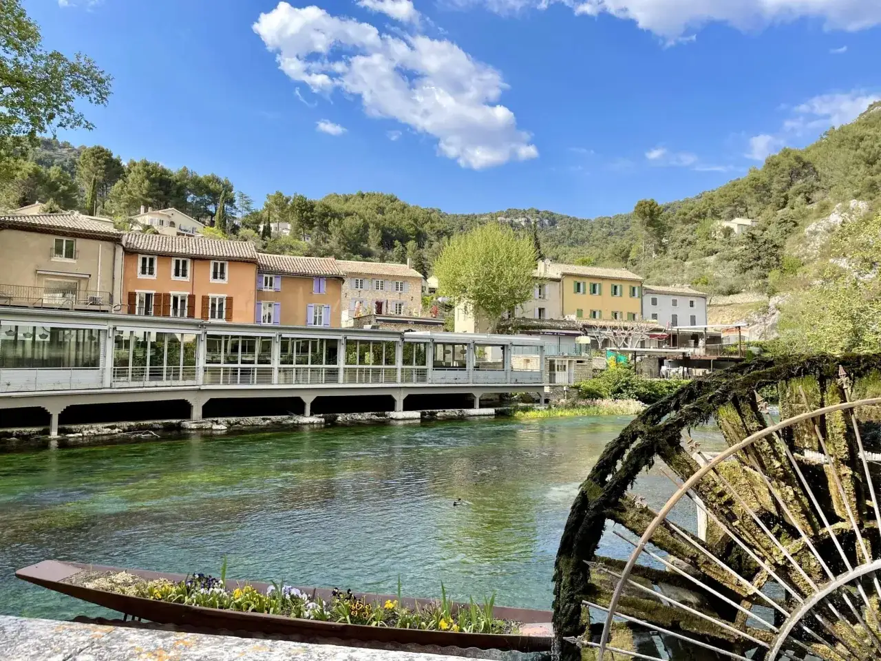 Le **gouffre de Fontaine-de-Vaucluse** vu depuis la rivière, avec une roue à aubes ancienne et des maisons colorées en arrière-plan.