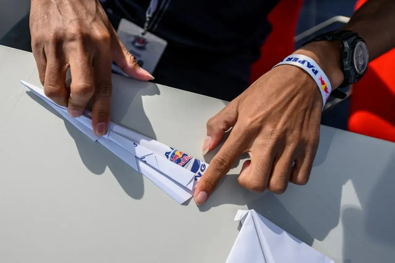 Instrukcja jak zrobić samolot z papieru. Dłonie składają biały papier, tworząc skrzydła. Na papierze widoczne logo Red Bull.