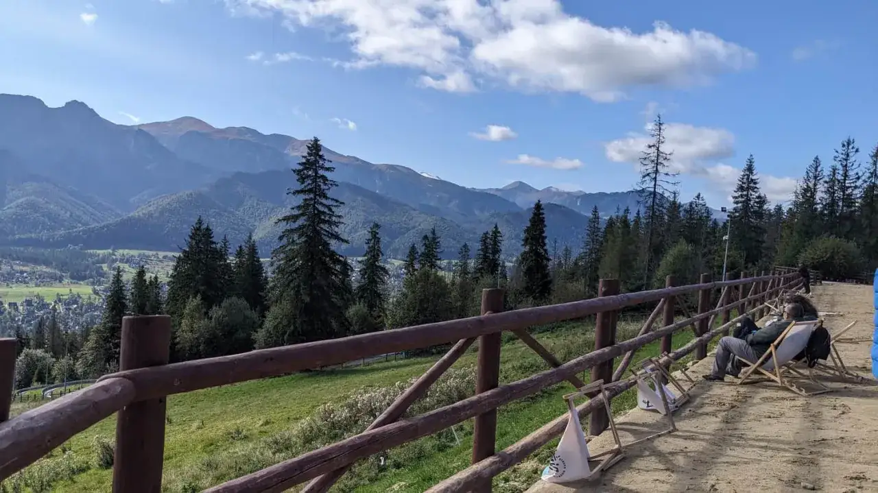 Widok na Tatry z Gubał&oacute;wki. Niebieski szlak prowadzi przez las, a na pierwszym planie drewniane ogrodzenie i odpoczywający turysta.