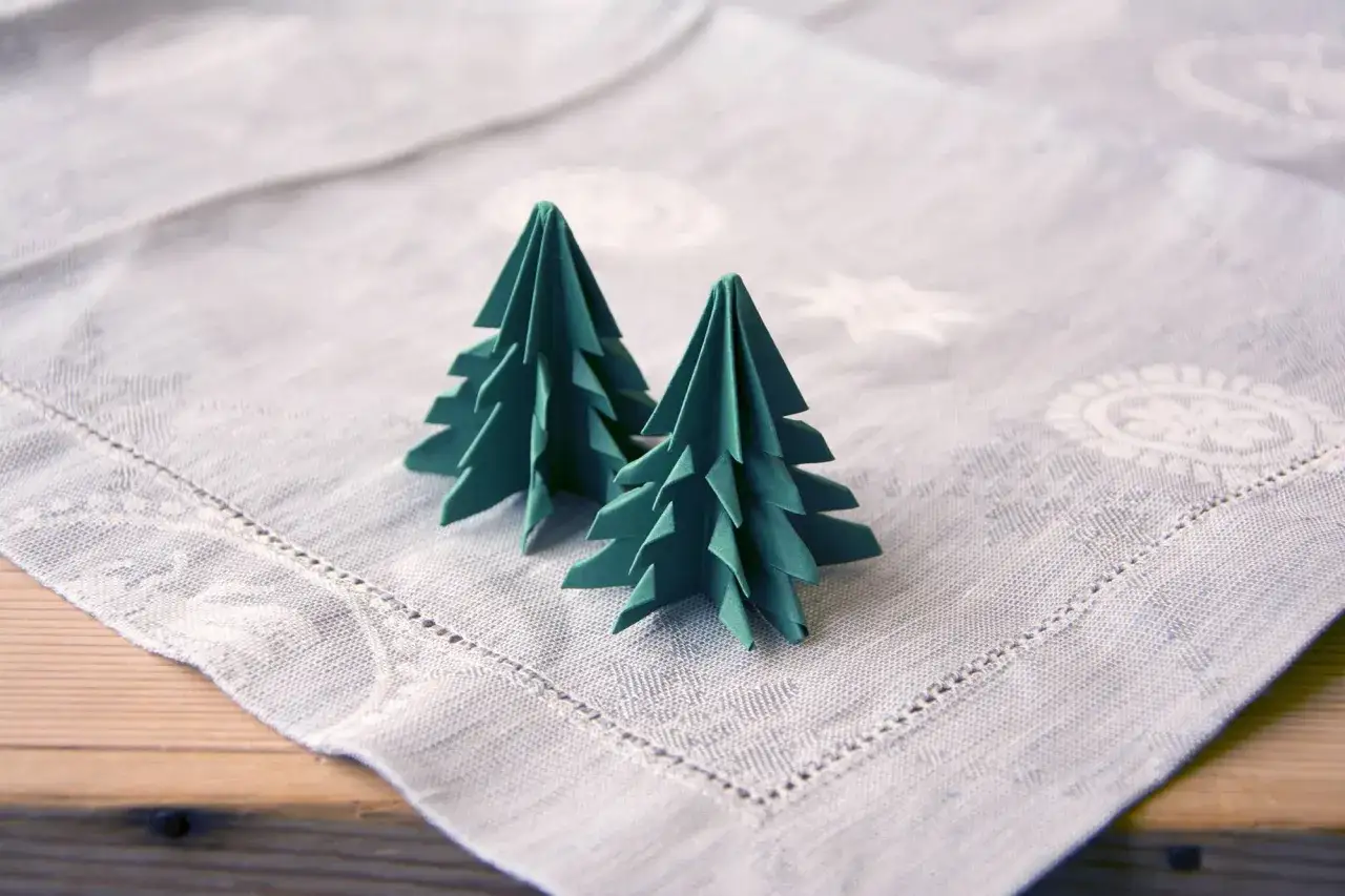 Zwei gr&uuml;ne Origami-Tannenb&auml;ume, die auf einer Tischdecke stehen. Eine einfache tannenbaum falten anleitung f&uuml;r Weihnachtsdekoration.