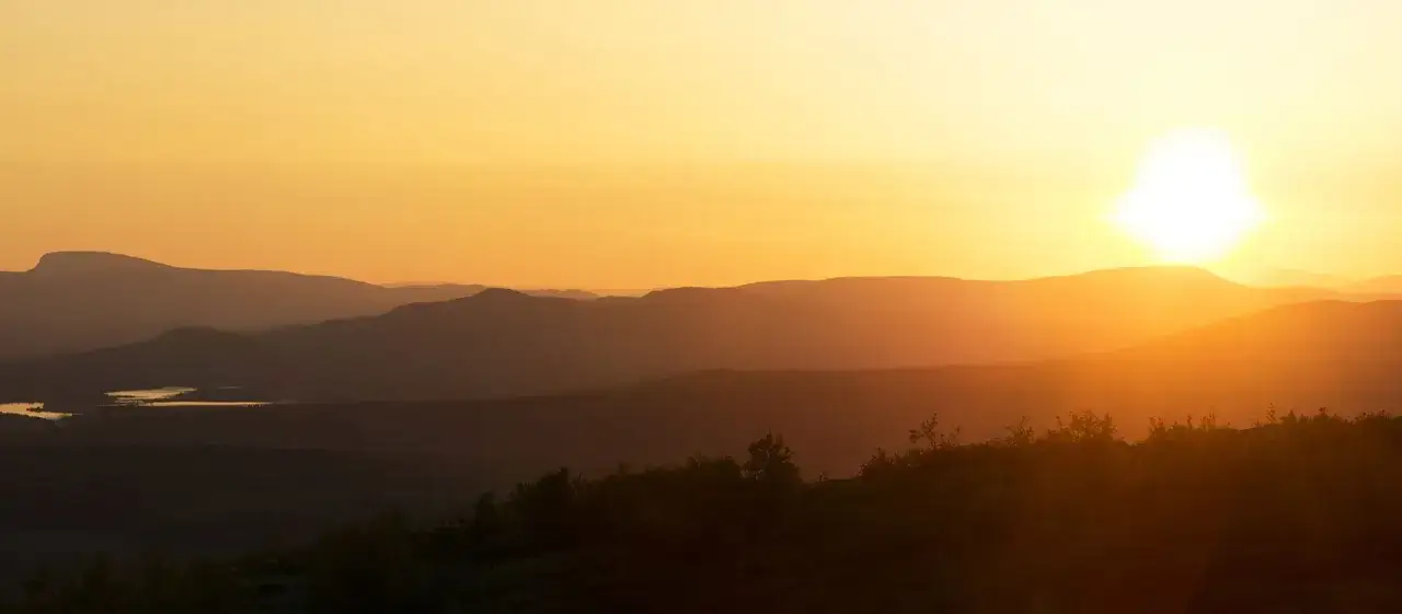 Solnedgång över Lapplands natur. Bergskedjor i silhuett mot en gyllene himmel. En sjö glimmar i fjärran.