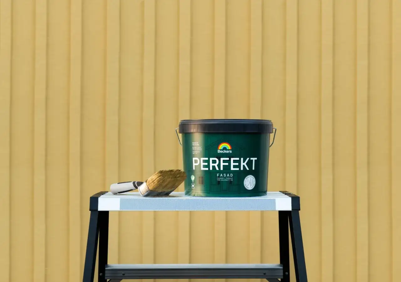 En burk Beckers Perfekt fasadf&auml;rg och en pensel st&aring;r p&aring; en stege framf&ouml;r en gul tr&auml;fasad. Perfekt f&ouml;r att m&aring;la utomhus, &auml;ven vid varierande luftfuktighet.