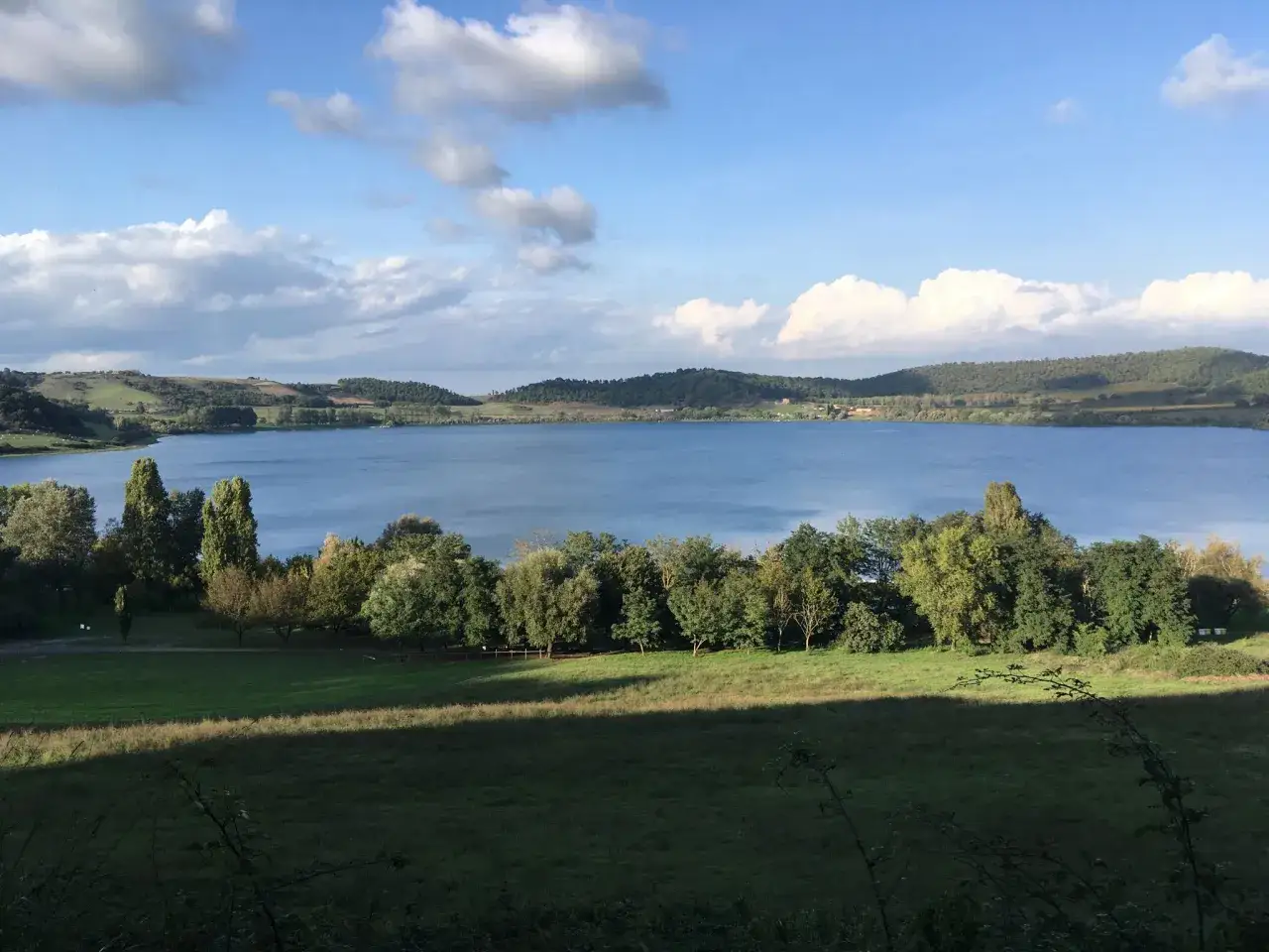 Vasta distesa azzurra del lago di Martignano, circondata da dolci colline verdi e alberi rigogliosi sotto un cielo con nuvole bianche.