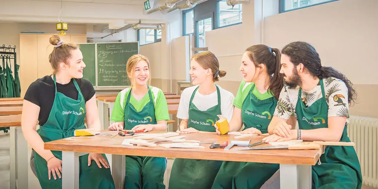 Fünf junge Leute in grünen Schürzen lernen an Holzarbeiten. Die Ausbildung Ergotherapie kostenfrei ist hier möglich.