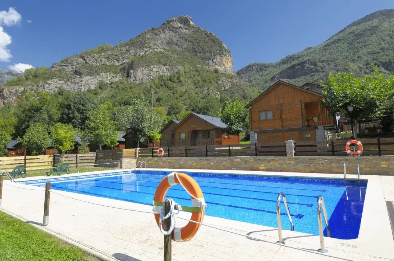 Piscina azul brillante en un entorno montañoso, rodeada de cabañas de madera. Un lugar ideal para disfrutar de los mejores campings de Catalunya.