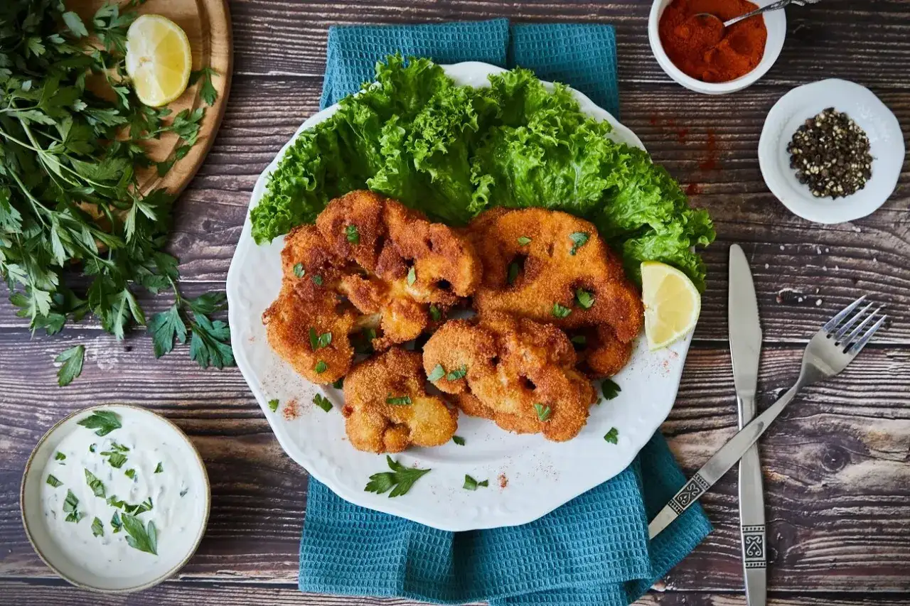 Knuspriges Blumenkohl Schnitzel auf einem Teller mit Salat, Zitrone und Dip. Dazu Paprikapulver und Pfefferkörner.