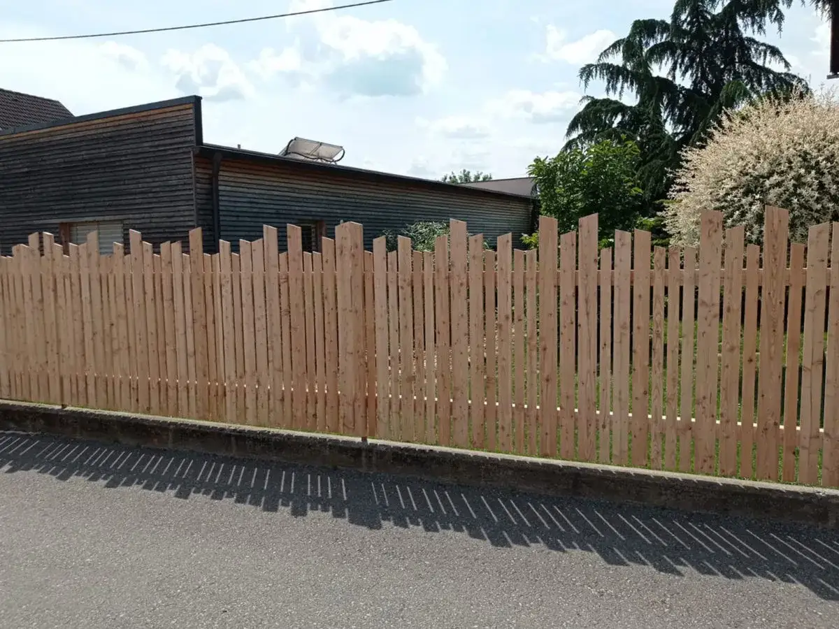 Ein neuer, heller Holzzaun, der zeigt, wie man einen zaun selber bauen billig kann. Dahinter ein Haus und B&auml;ume.