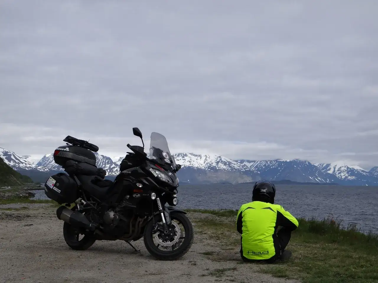 Motocyklista w jaskrawej kurtce siedzi nad wodą, podziwiając ośnieżone g&oacute;ry. Nordkapp pogoda jest typowo zmienna, chmury wiszą nisko.