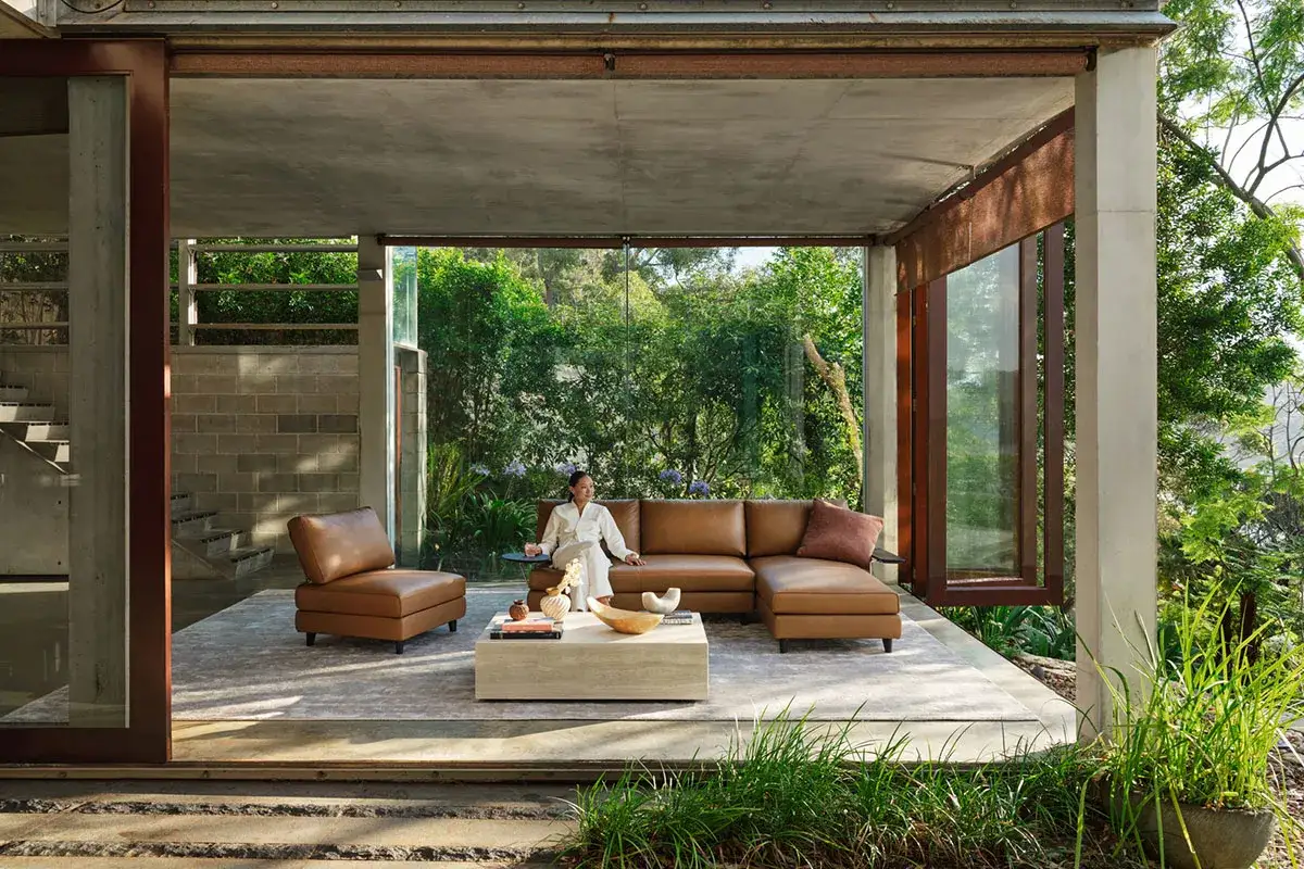 Un salon ouvert sur la nature, avec un canapé en cuir marron et une femme assise. Ce logement du futur s'intègre parfaitement à son environnement verdoyant.