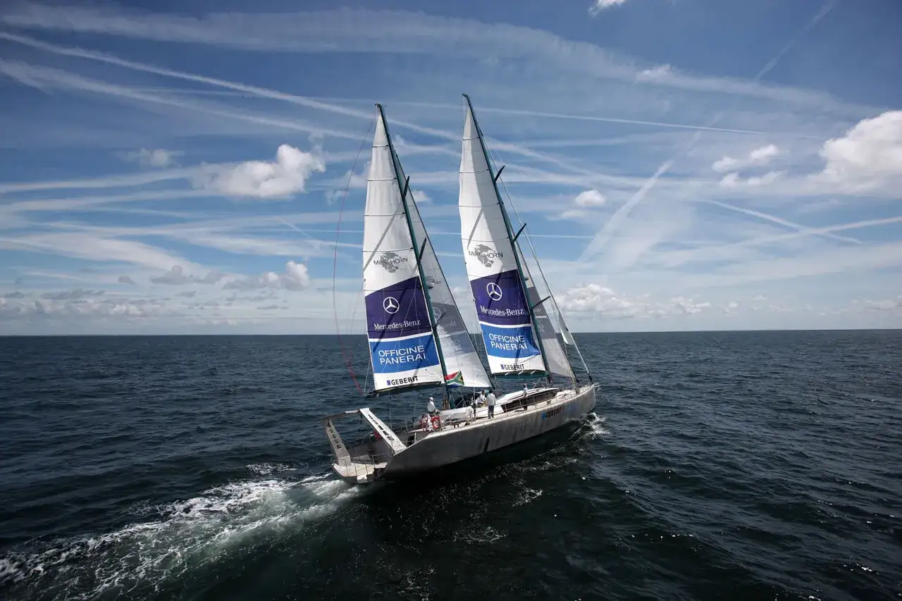 Le voilier Pangaea de Mike Horn navigue sur une mer calme sous un ciel bleu parsemé de nuages.