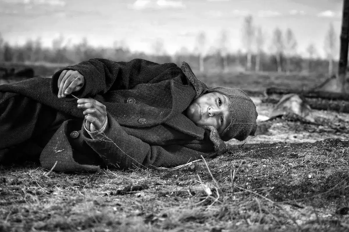 Chłopiec w płaszczu i czapce leży na ziemi, trzymając patyk. Scena przypomina kadr z filmu