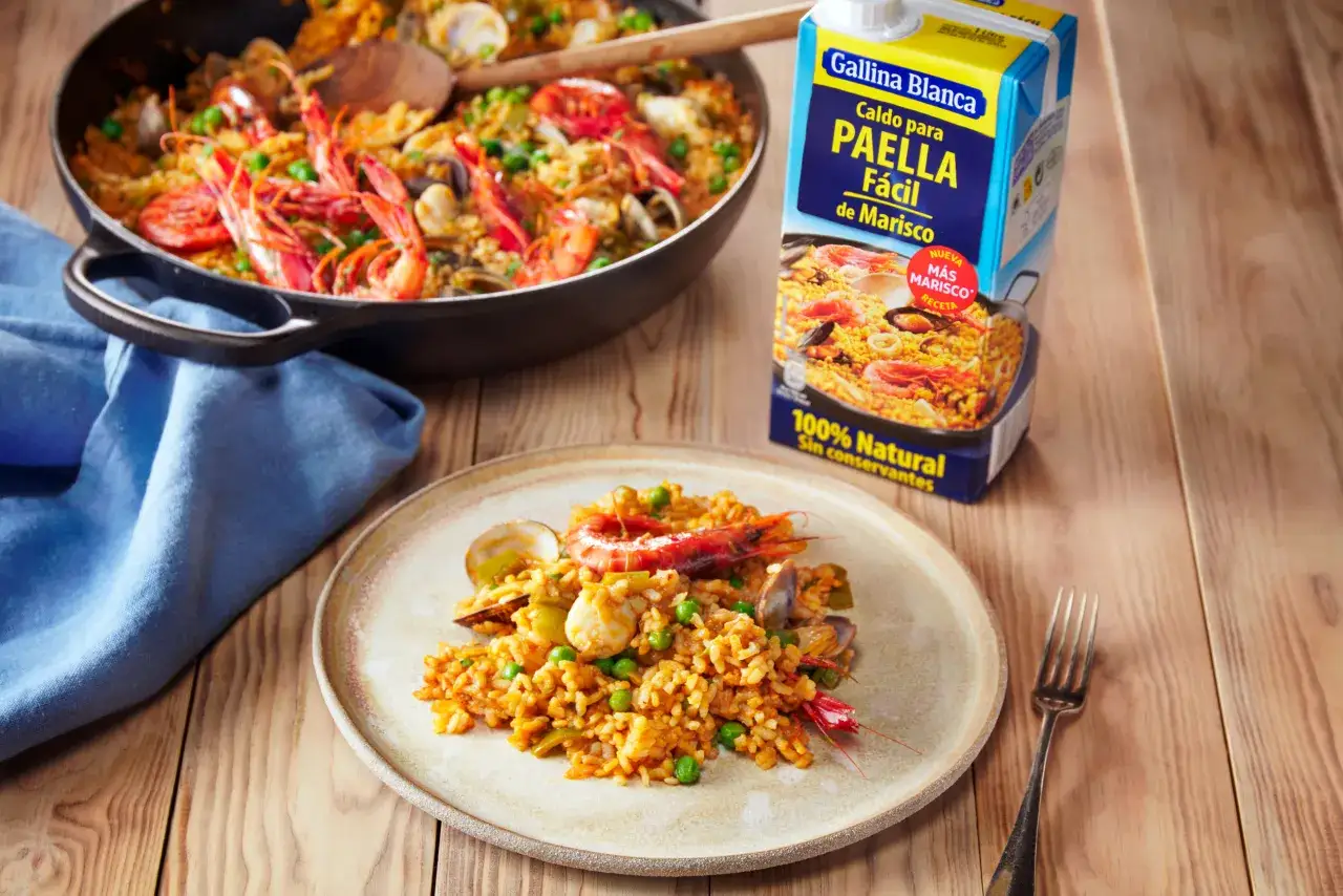 Deliciosa paella de marisco servida en plato y paellera, con un cart&oacute;n de caldo Gallina Blanca al lado.