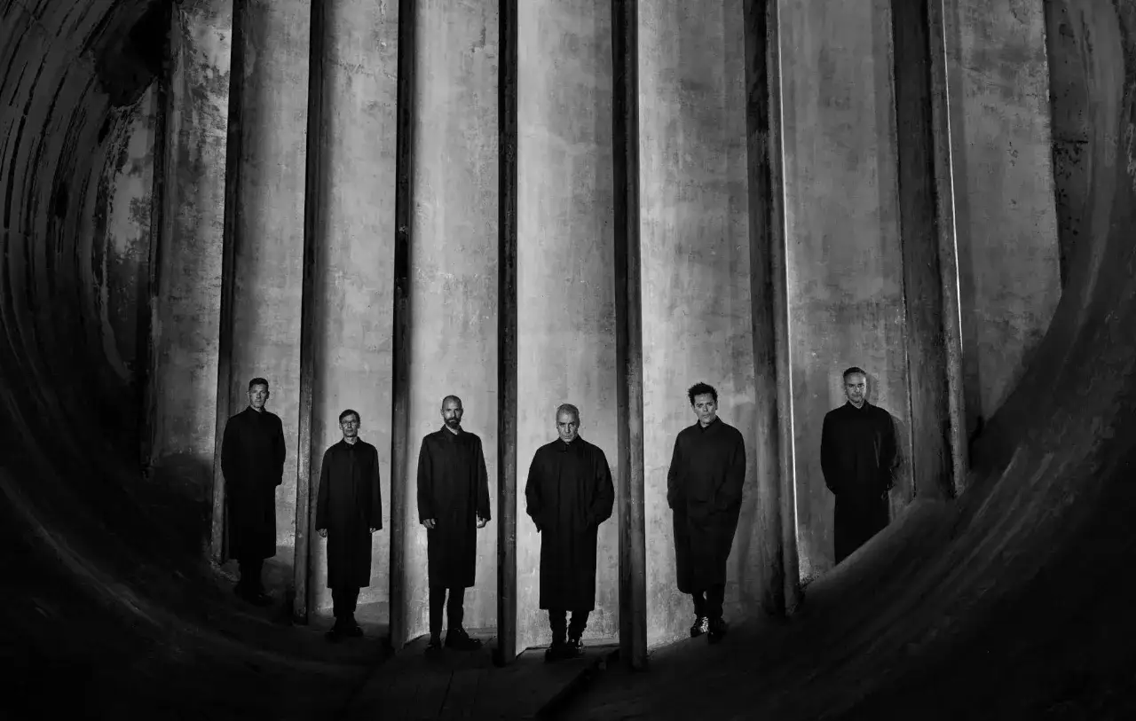 Siedmiu mężczyzn w długich płaszczach, jakby z okładki Rammstein Zeit, stoi w industrialnym wnętrzu.