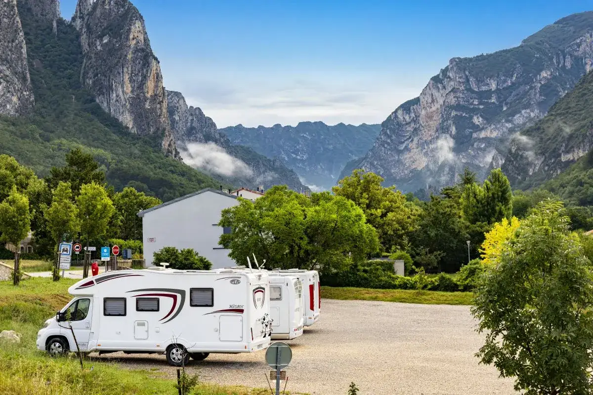 Trois camping-cars garés dans un parking rocailleux, avec des montagnes imposantes en arrière-plan. Un panneau de signalisation indique un 