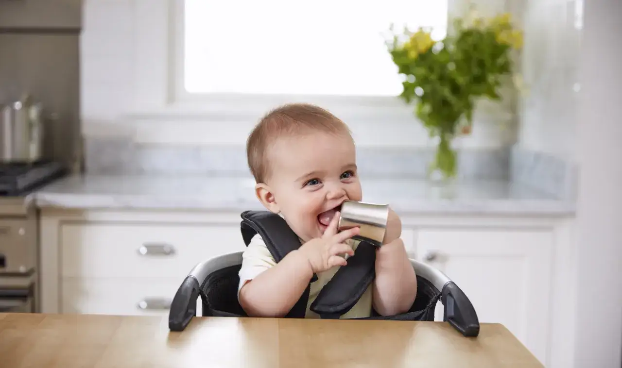 Ein fröhliches Baby im Hochstuhl, das neugierig einen Becher erkundet. Wann darf ein Baby Wasser trinken?