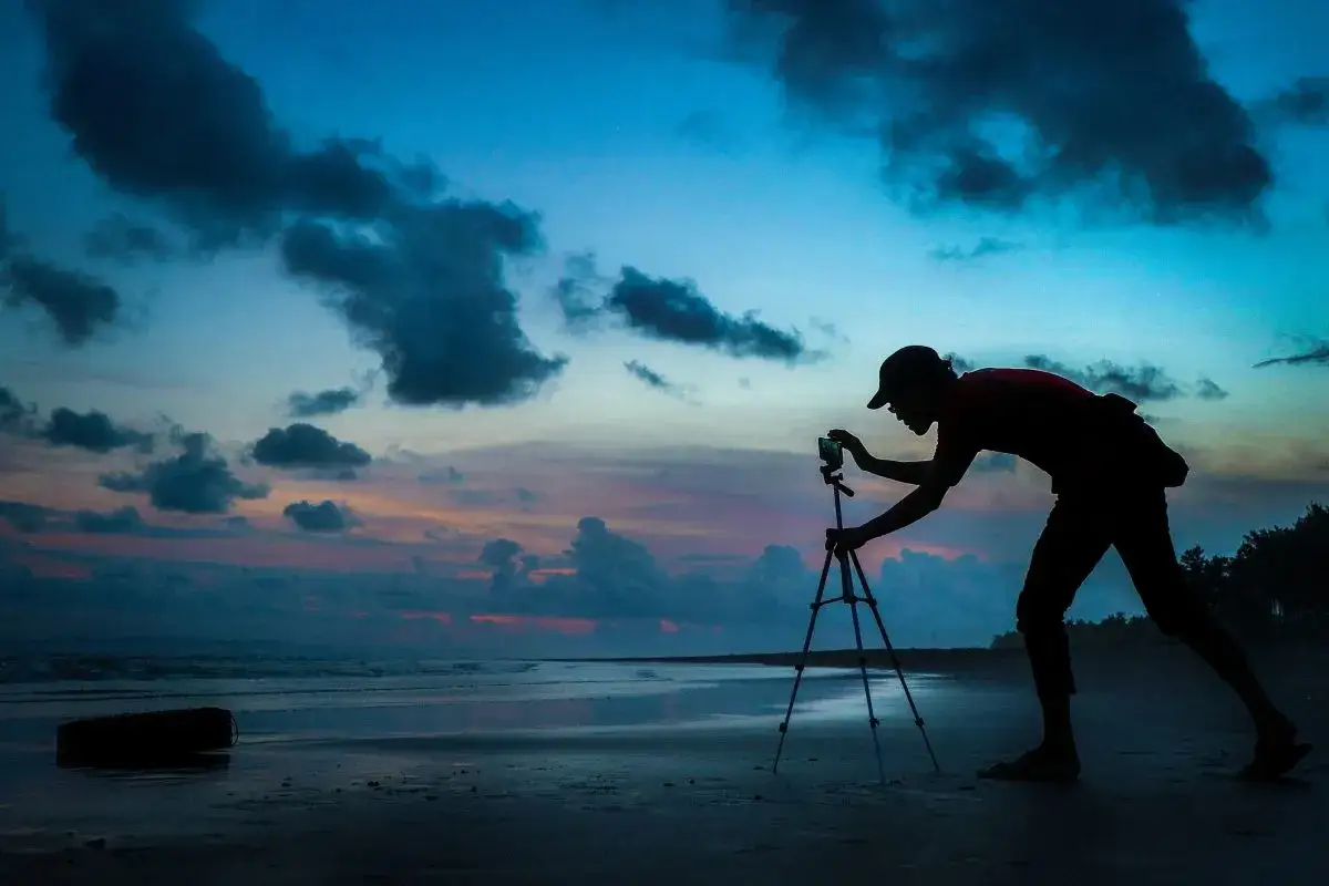 Sylwetka fotografa na plaży, ustawiająca telefon na statywie. Chce zrobić zdjęcie gwiazd, ale niebo jest jeszcze jasne.